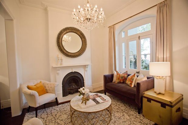 Elegant traditional living room with ornate ceiling cornices, a white mantel fireplace, arched French doors, long drapery, crystal chandelier, round decorative mirror, patterned rug, upholstered armchair, burgundy sofa, and refined brass-toned side table.