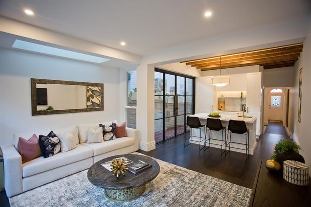 A bright, open-plan living and kitchen area with a white sectional sofa, patterned area rug, round coffee table, and large wall mirror in the foreground. The adjoining kitchen has a white island with three bar stools, integrated cabinetry, pendant lighting, and dark flooring. Black-framed glazed doors open to a balcony or exterior view, while exposed timber ceiling beams add warmth and character.