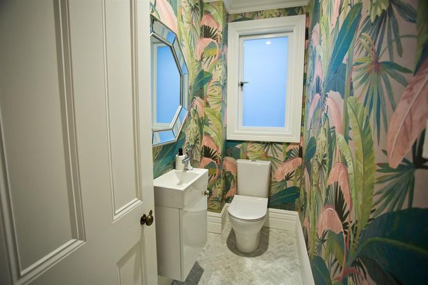 Compact powder room with a white pedestal-style vanity and toilet, a frosted window, decorative framed mirror, and vivid tropical botanical wallpaper covering the walls. The floor has a pale herringbone-patterned finish and the white paneled door is partially open.