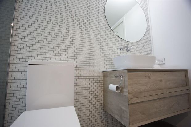 A compact contemporary bathroom with a wall-mounted timber-look vanity, white vessel basin, chrome wall-mounted tapware, round mirror, wall-hung toilet, and small light grey brick-pattern wall tiles. The palette is predominantly white, light grey, and light wood.