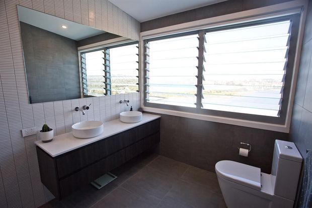 Contemporary bathroom with a double floating dark-wood vanity, two vessel basins, wall-mounted faucets, a large mirrored cabinet, textured vertical wall tiling, and a toilet. Wide louvred windows provide abundant daylight and views beyond.