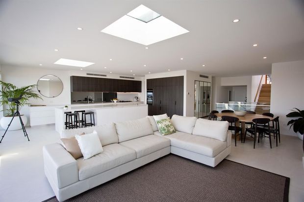 Bright open-plan contemporary interior combining a living room, kitchen and dining area. A large white sectional sofa faces the camera, with a dark textured rug beneath. The kitchen features a white island with bar seating, dark timber cabinetry and integrated appliances. A glass dining table sits beside a staircase, while skylights and recessed lighting provide abundant illumination.