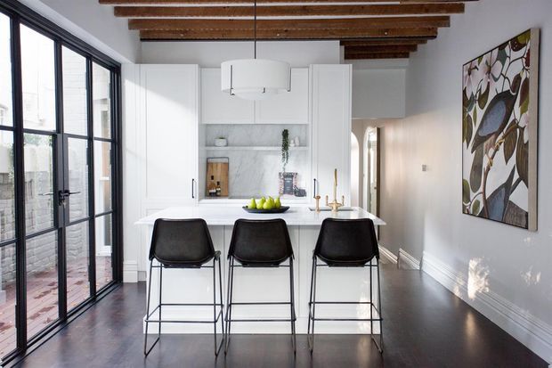 Bright, refined kitchen interior with a white island and three black bar stools, white shaker-style cabinetry, marble-look backsplash and benchtop, dark timber flooring, black-framed glazed doors, and exposed timber ceiling beams. A large botanical artwork adds visual interest to the otherwise neutral scheme.