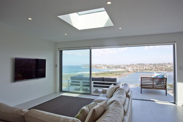 Bright contemporary living room with a large sectional sofa, wall-mounted television, tiled floor, and expansive sliding glass doors opening to a balcony with a waterfront view. A skylight and recessed ceiling lights provide additional natural and artificial illumination.