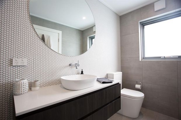 Contemporary bathroom featuring a floating dark-wood vanity with a white engineered-stone benchtop, round vessel basin, wall-mounted tapware, large circular mirror, white toilet, and grey tiled walls. A small hexagonal mosaic-tile feature wall contrasts with larger grey wall and floor tiles, while a high horizontal window provides natural light.