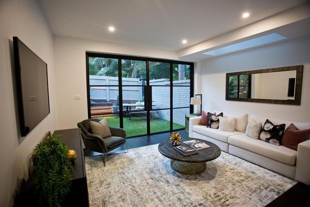 Contemporary living room with a white sectional sofa, dark round coffee table, armchair, large area rug, wall-mounted television, and a full-height black-framed glazed opening to a small outdoor area. Recessed ceiling lights and a linear skylight provide a bright, refined interior.