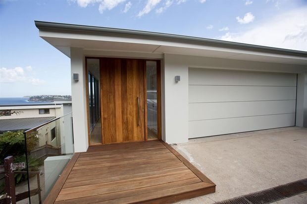 A contemporary residential entry facade viewed from outside, featuring a broad low-pitched roof, white rendered walls, a large warm timber pivot door with narrow glazed sidelights, and an integrated horizontal-panel garage door. A timber deck entry platform and concrete driveway occupy the foreground, with a distant coastal or suburban outlook visible to the left.