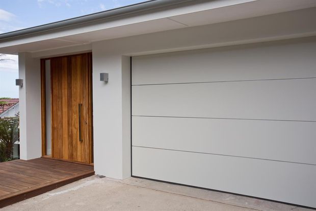 Contemporary residential facade showing a large white sectional garage door beside a tall timber entry door. The entrance sits beneath a broad flat roof overhang, with a timber-clad porch deck and minimalist white rendered walls.