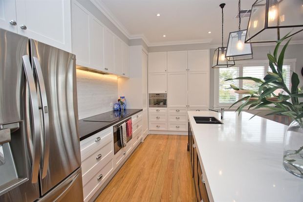 Bright, spacious contemporary kitchen with white shaker-style cabinetry, stainless-steel French-door refrigerator, dark benchtops, a large white island, timber flooring, integrated appliances, and prominent black-framed pendant lights. A window provides natural light, while a leafy plant adds a small decorative accent.