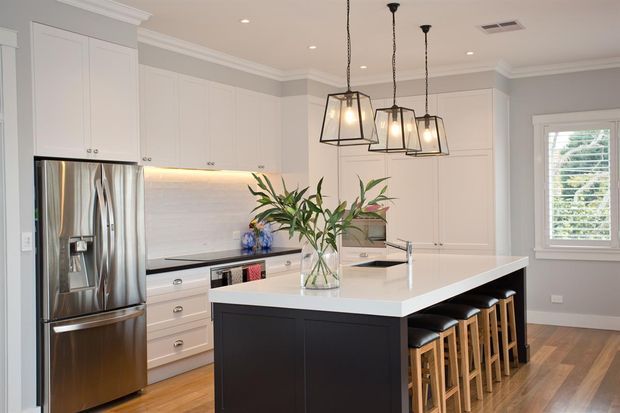 Bright, refined transitional kitchen with white shaker-style cabinetry, a dark island base, white stone benchtops, stainless-steel appliances, a pale tiled backsplash, timber flooring, and three black-framed pendant lights. The island includes seating for five or more, and a window with plantation shutters brings in natural light.