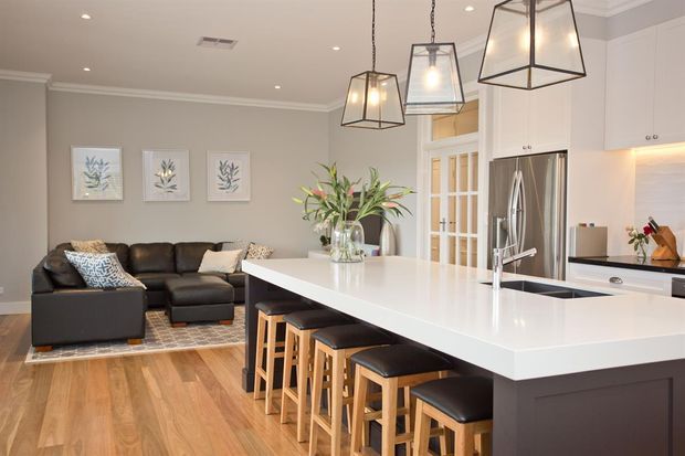 A bright, open-plan kitchen and living area with a large white island, dark base cabinetry, timber bar stools, stainless-steel appliances, and pendant lighting. The adjoining living room includes a dark sectional sofa, patterned rug, and botanical wall art. Neutral grey walls, white cabinetry, and warm timber flooring create a refined contemporary family space.