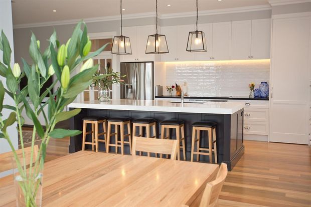 Bright, spacious kitchen and dining area with a large dark island, timber bar stools, pale cabinetry, white tiled backsplash, pendant lights, and a timber dining table in the foreground. Indoor plants add a natural accent, while decorative wall mouldings and a coffered-style ceiling contribute a refined traditional character.