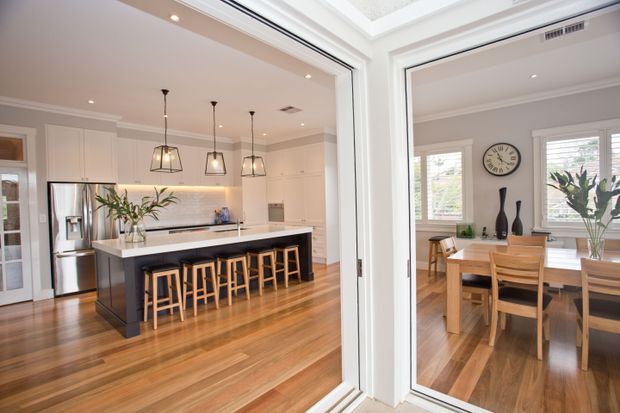 Bright open-plan view through internal glazing, showing a contemporary kitchen with a large dark island and timber stools on the left, and an adjoining dining room with a timber table and chairs on the right. The space features warm timber flooring, white cabinetry, pale grey walls, white trim, pendant lighting, and abundant natural light.