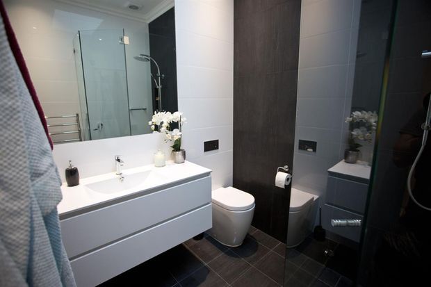 Modern bathroom with a wall-mounted double-drawer vanity, white basin, large mirror, glass shower enclosure, toilet, and dark tiled feature wall and floor. The space has a clean, contemporary monochrome palette with white surfaces contrasted by charcoal and dark wood-look finishes.