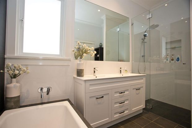 Bright, refined contemporary bathroom with a freestanding bathtub, white vanity with drawers, large mirror, and frameless glass walk-in shower. White wall tiles, dark floor tiles, recessed lighting, and simple floral styling create a clean high-contrast scheme.