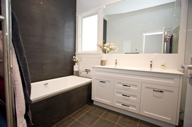 Bright contemporary bathroom featuring a white bathtub set into dark charcoal tiled surrounds, matching dark floor tiles, and a large white vanity with multiple drawers and integrated basins beneath a wide wall mirror. A window, recessed ceiling lights, and minimal floral styling complete the clean, monochromatic space.