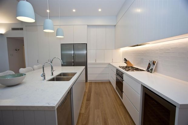 Bright contemporary kitchen with a large white island featuring a double stainless-steel sink and curved tapware, white handleless cabinetry, integrated stainless-steel refrigerator, gas cooktop, and white subway-tile splashback. Light timber flooring and pale blue pendant lights add warmth and colour contrast.