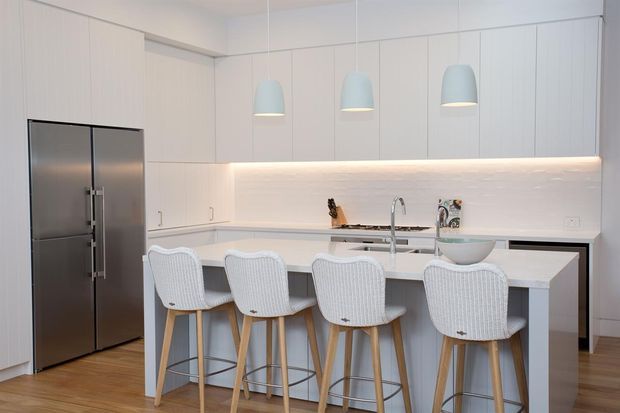 Bright, contemporary white kitchen with extensive flat-panel cabinetry, a large central island with seating for four, stainless-steel French-door refrigerator, integrated cooktop and sink, illuminated backsplash, and three pale pendant lights. Light timber flooring and woven bar stools add warmth and texture.
