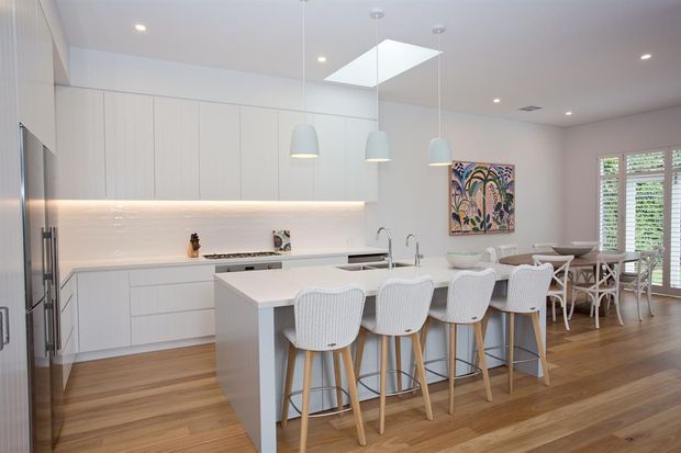 Bright open-plan kitchen and dining area with white flat-panel cabinetry, a large white island with four stools, pendant lights, skylight, timber flooring, and a dining table beside louvered glazing. The space has a clean, contemporary coastal feel with colourful artwork adding a small accent.