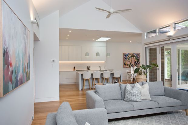 Bright open-plan living area with a large grey sectional sofa in the foreground and a contemporary white kitchen with a central island and bar seating beyond. The space features warm timber flooring, white cabinetry, recessed lighting, a skylight, clerestory windows, extensive glazed doors, a ceiling fan, and colourful artwork.