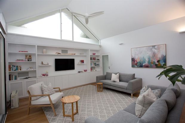 Bright contemporary living room with a vaulted ceiling and prominent triangular clerestory glazing. Built-in white shelving and cabinetry surround a wall-mounted television, while grey sofas, a patterned rug, timber occasional tables, and a large abstract artwork create a comfortable family seating area. A ceiling fan and indoor plant complete the space.
