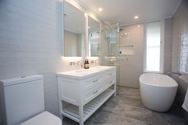 Bright contemporary bathroom with a white double-sink vanity, wall-mounted mirrors, glass-enclosed shower, freestanding oval bathtub, toilet, and pale textured wall tiles. The floor has a grey stone-look finish, with recessed ceiling lighting and a narrow frosted window.