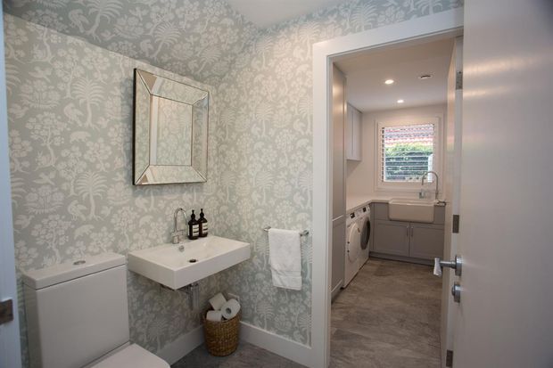 A compact powder room with pale botanical patterned wallpaper, a wall-mounted white basin, toilet, decorative framed mirror, and towel rail. An open doorway reveals an adjoining laundry with cabinetry, washing machine, dryer, utility sink, and a window with blinds. The room has a refined traditional interior with soft neutral tones and patterned surfaces.