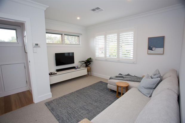Bright, compact living room with a large light-grey sectional sofa, textured grey area rug, white TV console and wall-mounted television. The room has white walls, decorative cornices, plantation shutters, recessed lighting, and a small timber side table. A partially visible entry door and timber-floored adjoining area appear at left.