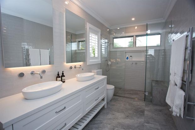 Bright, spacious contemporary bathroom with a long white double vanity, two vessel basins, wall-mounted taps, large mirrored cabinetry, grey tiled walls and floor, and a frameless glass walk-in shower. White plantation shutters, recessed ceiling lights, a heated towel rail, and a toilet complete the room.