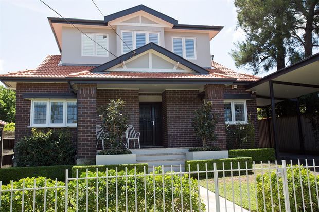 Street-facing view of a two-storey suburban brick house with a prominent front porch, dark brick walls, white-framed windows with shutters, a terracotta tiled gabled roof, and a covered carport to the right. A white metal front fence, hedges, paving, and low-maintenance landscaping frame the entrance.