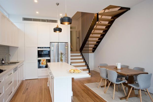Bright open-plan kitchen and dining area with white shaker-style cabinetry, a central island, integrated appliances, timber flooring and a timber dining table. A striking open-riser staircase with dark structural framing and timber treads rises beside the kitchen, with vertical timber screening at the upper landing. Contemporary pendant lighting and neutral furnishings complete the space.