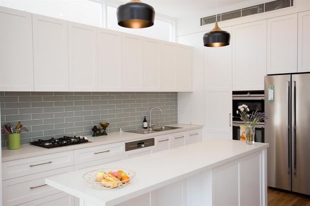 Bright, spacious contemporary kitchen with white shaker-style cabinetry, a large white island, grey subway-tile splashback, stainless-steel appliances, timber flooring, and black-and-brass pendant lights. The layout includes extensive overhead storage, integrated cooking appliances, a sink run, and a generous island bench.