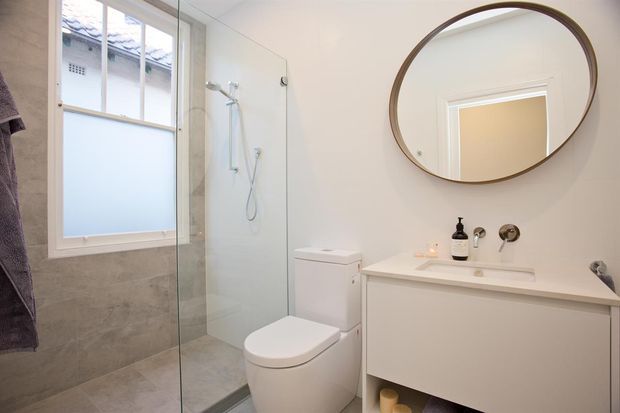 Bright contemporary bathroom with a frameless glass walk-in shower, wall-mounted shower fittings, large frosted window, tiled lower walls and floor, toilet, and a floating white vanity with integrated basin. A large round timber-toned mirror and brushed metal tapware add understated warmth.