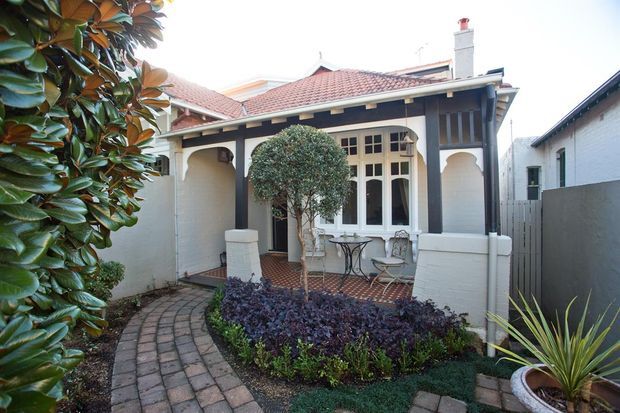 Exterior view of a compact heritage-style residence with white rendered walls, dark timber framing, a red terracotta tiled roof, decorative arched porch openings, and multi-pane windows. A curved brick-paved path leads through a landscaped front garden with clipped shrubs and ornamental planting.