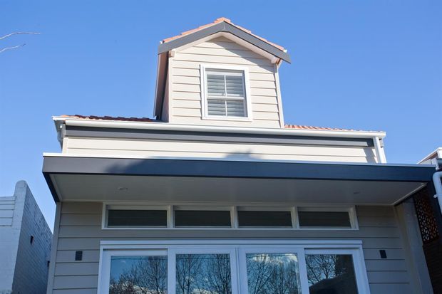 Exterior upward view of a narrow multi-storey residential building with a prominent upper dormer or gabled room, horizontal light-coloured weatherboard cladding, white-framed windows and doors, and a low-pitched tiled roof. The facade combines traditional detailing with a contemporary lower-level awning and glazing.