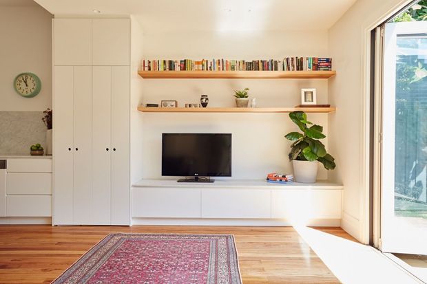 Bright, compact contemporary living room with warm timber flooring, white built-in cabinetry, open display shelves, a wall-mounted television, indoor plant, and a patterned area rug. Full-height glazed doors open to an outdoor area, bringing in abundant natural light.
