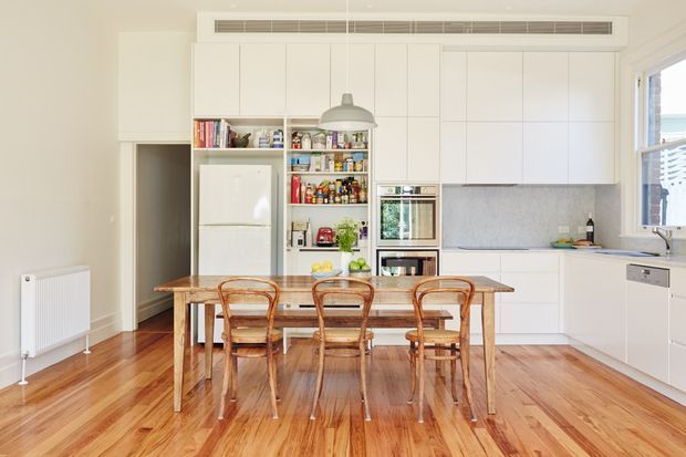 Bright, open-plan kitchen and dining area with white full-height cabinetry, integrated appliances, open shelving, a pale stone or concrete-look backsplash, and a timber dining table surrounded by bentwood chairs. Warm polished timber flooring contrasts with the crisp white joinery, while a large window brings in natural light. A wall-mounted split air-conditioning vent and radiator are also visible.