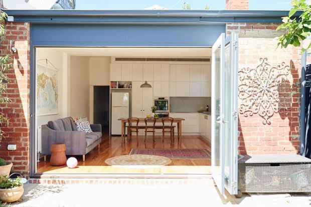Bright view from an outdoor terrace into a renovated brick home, showing a wide glazed opening with folded doors, a living room, and an open-plan white kitchen and dining area. The interior has polished timber flooring, classic timber dining furniture, a grey sofa, and a warm neutral palette. Exposed red brick walls and a blue-grey external frame provide architectural character.