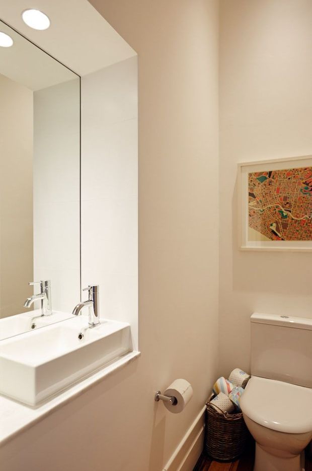 A compact powder room with a wall-mounted white basin, chrome faucet, large frameless mirror, toilet, recessed ceiling lights, beige painted walls, and white trim. A framed colourful artwork hangs above the toilet, with a woven basket beside it.