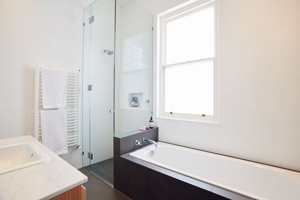 Bright contemporary bathroom featuring a freestanding bathtub beneath a large window, a glass-enclosed shower, wall-mounted towel radiator, and dark vanity cabinetry with a light stone benchtop. The room has a clean, neutral palette and abundant natural light.