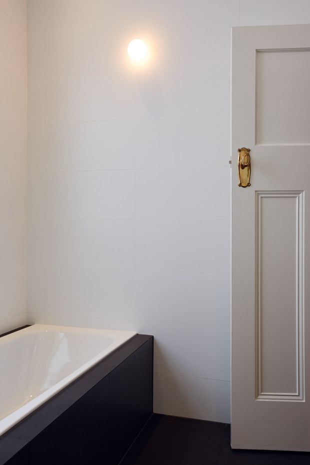A partial view of a compact, refined bathroom featuring a white built-in bathtub with dark cabinetry or paneling beneath, pale tiled walls, dark flooring, and a traditional raised-panel door with an ornate brass handle. A simple wall light provides warm illumination.