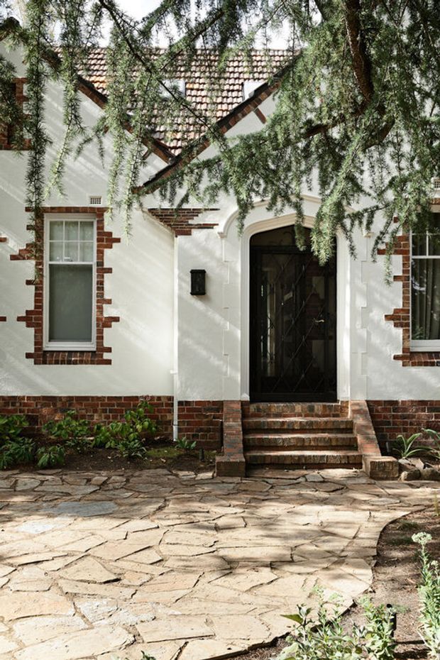 Front exterior view of a traditional brick-and-render home, partially framed by mature tree branches. The facade has white rendered walls, exposed red brick detailing around the windows and lower walls, a tiled pitched roof with intersecting gables, and a dark arched entry door reached by brick steps. An irregular natural-stone paved path and modest garden planting occupy the foreground.