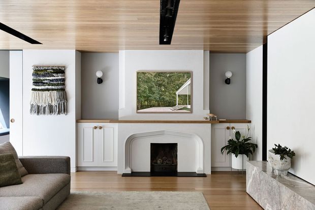 A refined contemporary living room centred on a white traditional-style fireplace with curved detailing, flanked by built-in cabinetry and a framed view of greenery. The room features a warm timber-lined ceiling, light timber flooring, white walls, dark linear ceiling lighting, neutral upholstered seating, and a pale stone media console with indoor plants.