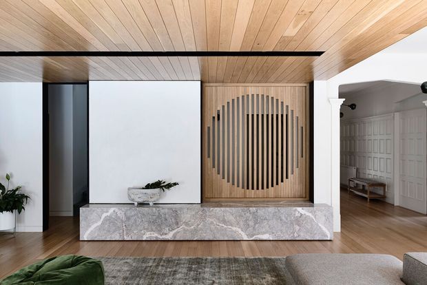 A refined contemporary living room featuring a full-height timber-lined ceiling and matching timber flooring. A large sculptural marble-clad built-in fireplace or media console anchors the room, with a geometric timber screen panel above it. White walls, dark-framed openings, understated furnishings, and a patterned rug create a warm, minimalist interior. An adjoining panelled room is visible through the arched opening.