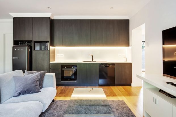Open-plan contemporary kitchen and living area with dark full-height cabinetry, integrated appliances, a pale stone or light benchtop, under-cabinet lighting, timber flooring, and a light sectional sofa in the foreground. White walls and ceiling cornices contrast with the dark joinery, while a wall-mounted television and low white media unit are visible to the right.