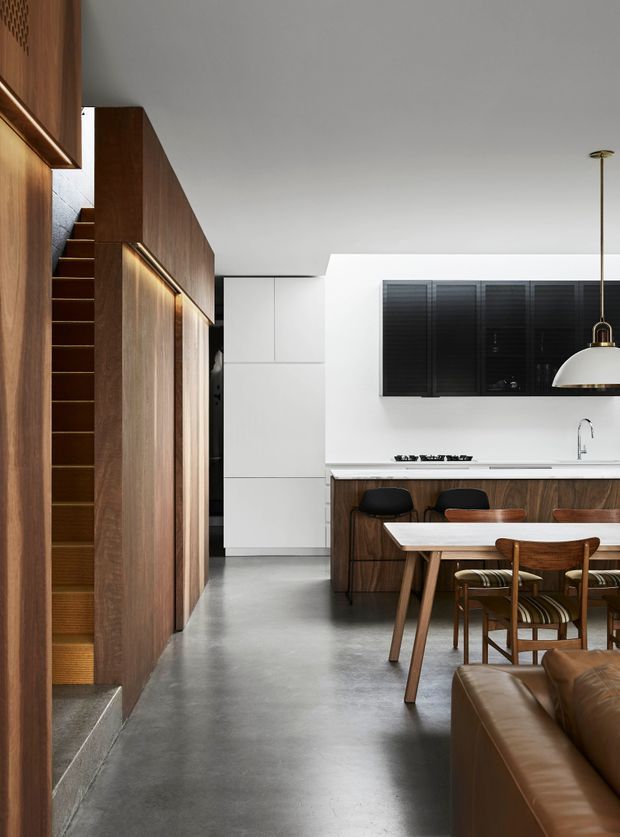 A refined open-plan interior showing a contemporary kitchen and dining area with a partial living room. The space features rich dark timber wall panelling and kitchen cabinetry, a white stone or solid-surface benchtop, integrated white cabinetry, polished grey concrete flooring, black window shutters, and a sculptural pendant light over a timber dining table. A timber-lined stairway and concealed storage wall add architectural interest.