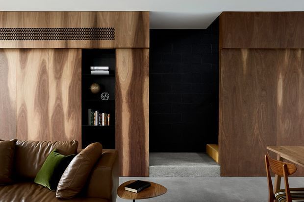 A richly detailed contemporary living room with extensive dark timber wall panelling and built-in joinery. A brown leather sofa with a green cushion sits beside a small round timber coffee table, while a black recessed shelving niche displays books and objects. A black brick-lined passage or feature wall leads to a raised area, and part of a timber dining table and chair is visible at right. The palette is warm timber, brown leather, black and grey concrete, with a refined, moody atmosphere.