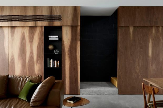A contemporary interior featuring extensive dark and mid-toned timber wall panelling and built-in cabinetry, with an integrated open bookshelf and perforated timber detail. A brown leather sofa with a green cushion sits beside a small round timber coffee table. A recessed black-painted brick passage or alcove leads to a raised grey-finished area, while part of a timber dining table and chairs is visible at right.