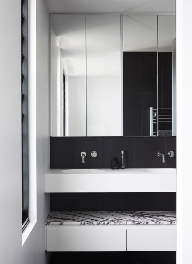 A compact contemporary bathroom vanity viewed straight on. The room features a wall-mounted white vanity with an integrated basin, black tiled backsplash and lower wall, mirrored wall cabinets, chrome tapware, and a striking white marble-look countertop or shelf with dark veining. A narrow window is visible along the left side, and a heated towel rail appears in the mirror reflection.