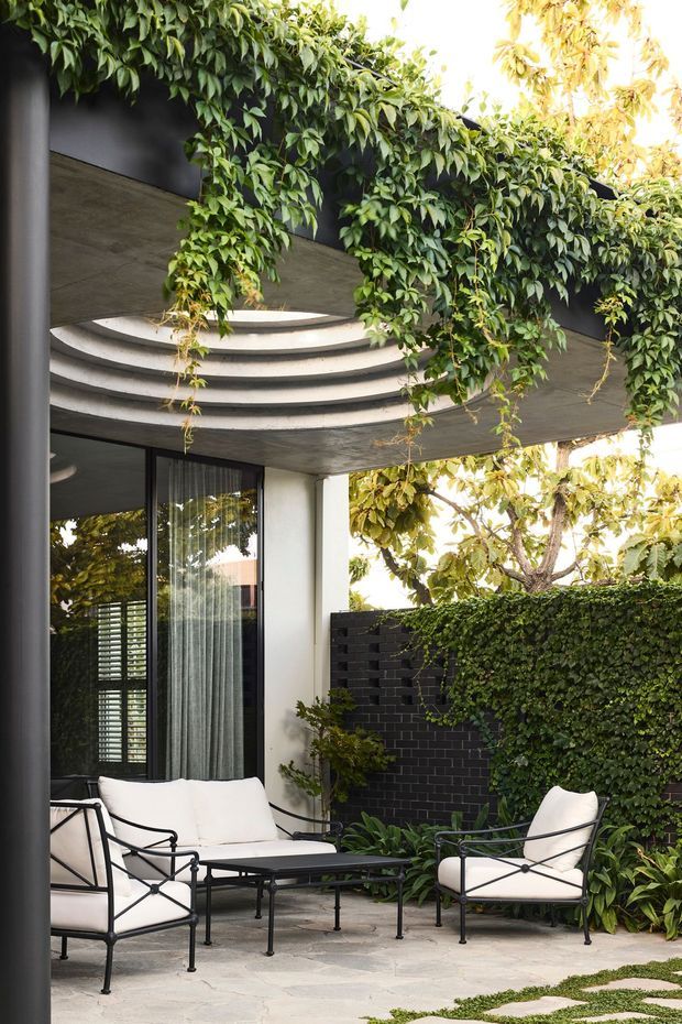 A refined outdoor courtyard lounge beneath a concrete overhang, with curved tiered concrete detailing and lush climbing greenery cascading across the structure. Black-framed glazed doors, a dark brick privacy wall, pale stone paving, and black metal lounge furniture with white cushions create a calm, garden-integrated setting.